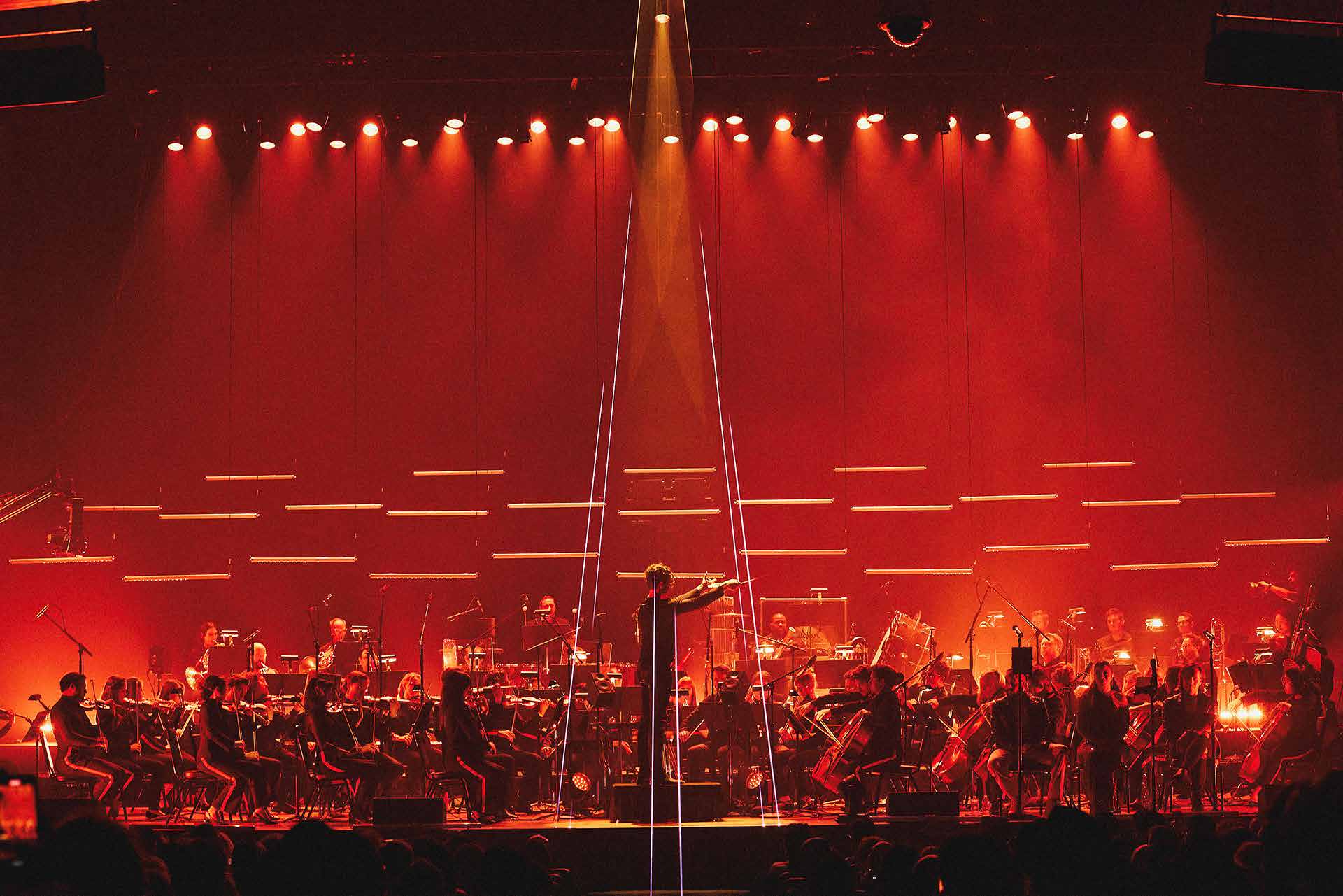 Full orchestra bathed in deep red stage lighting under a conductor